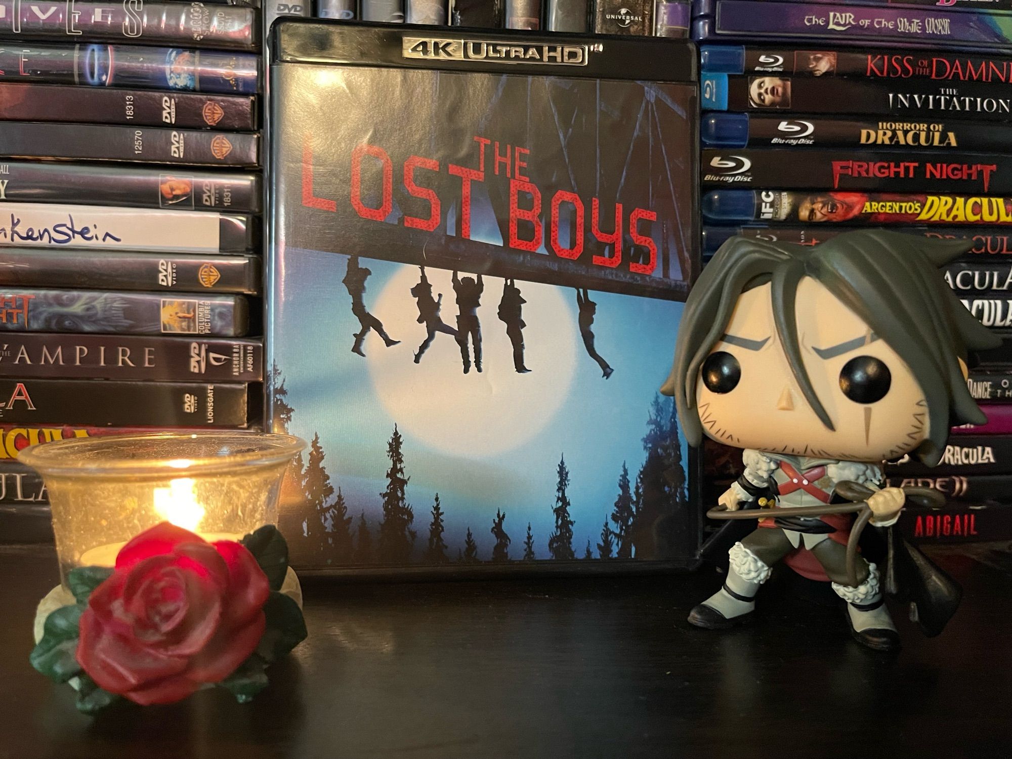 a copy of the blu ray of The Lost Boys sitting on a DVD shelf between a Funko Pop of Trevor Belmont from Castlevania and a lit tea light in a rose tea light holder.